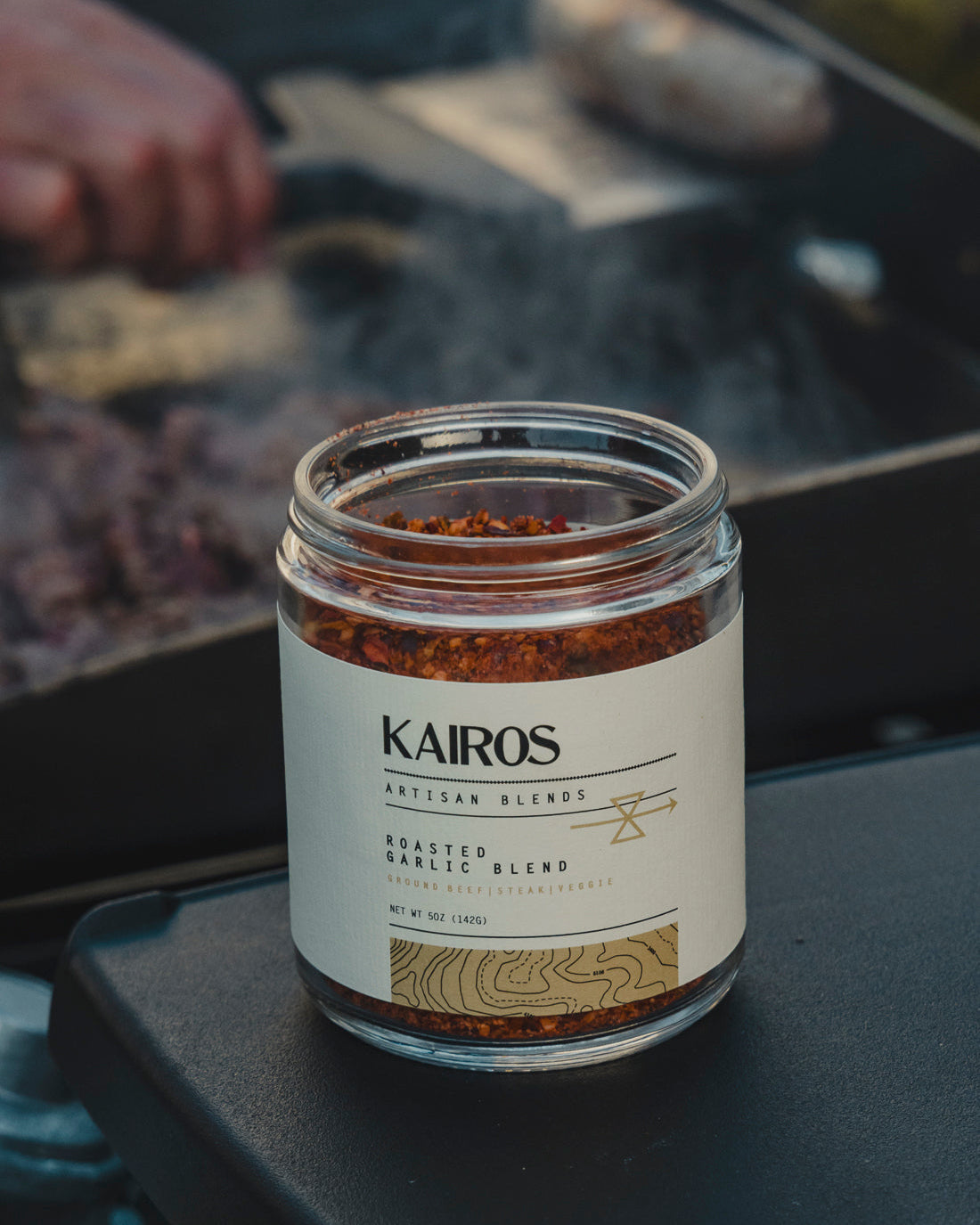 A glass jar of Kairos Natural Blends Roasted Garlic Blend seasoning, set on a dark surface with a blurred outdoor cooking scene in the background.