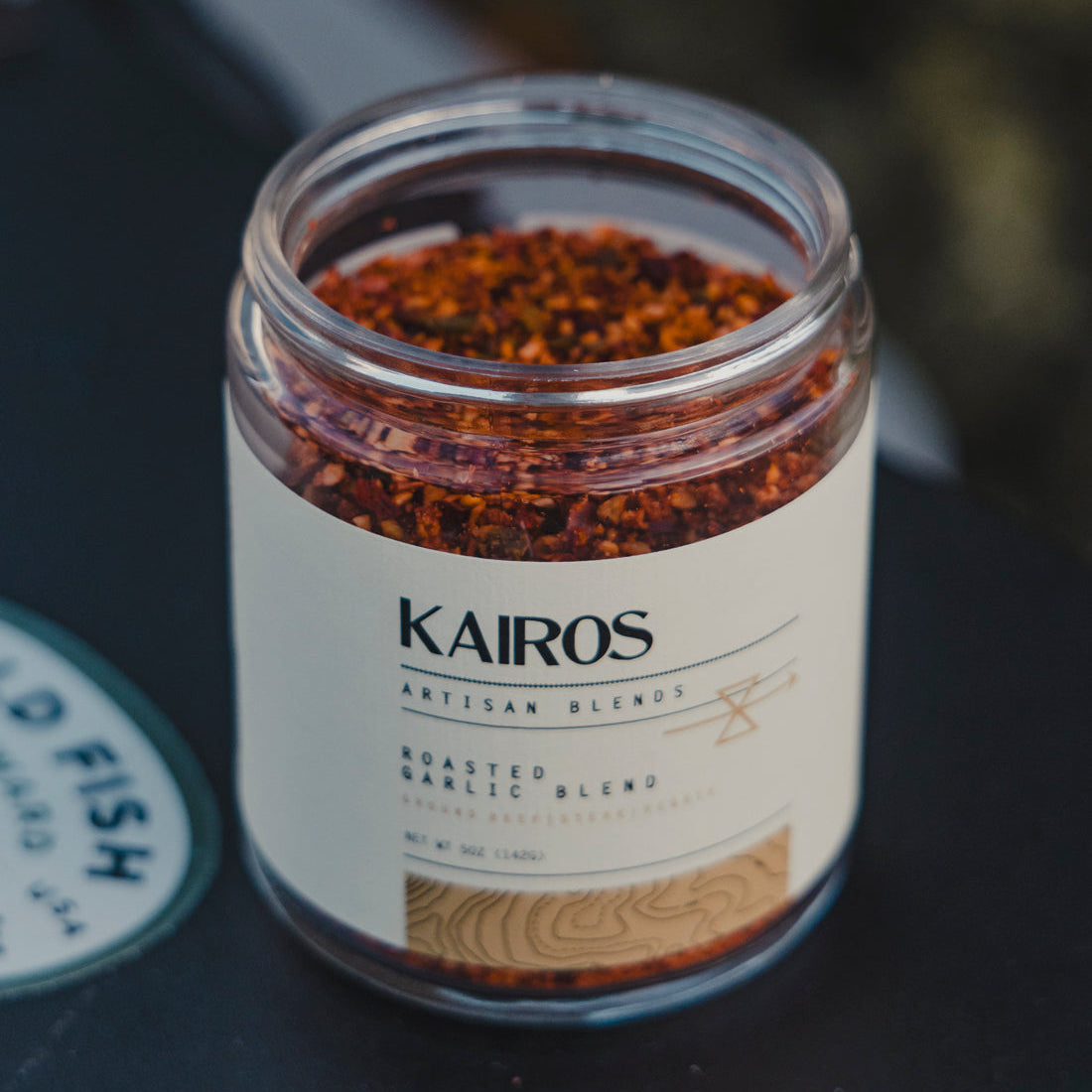 A clear glass jar of Kairos Artisan Blends Roasted Garlic Blend containing reddish-brown roasted garlic flakes, set on a dark surface with a blurred background.